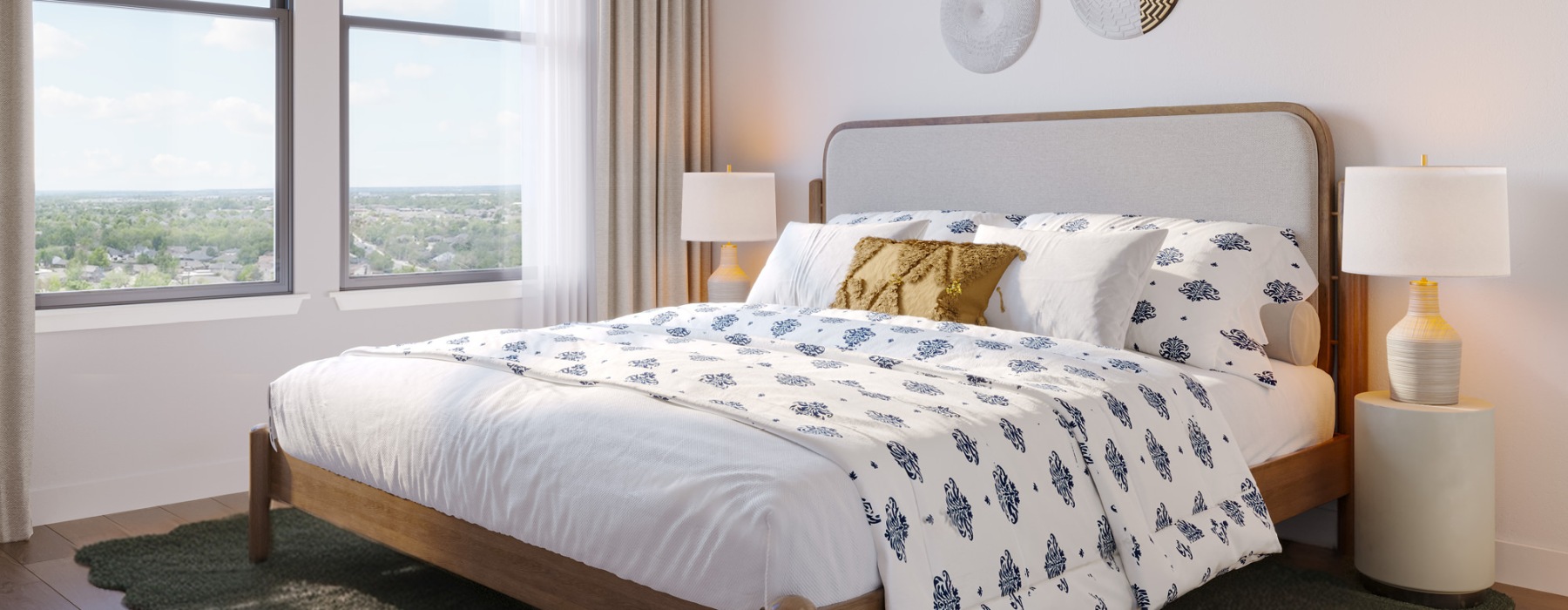 A bedroom at The Residences at OAK which features a large bed adorned with white and blue bedding, flanked by two nightstands each with a lamp. A window with curtains offers a view outside.